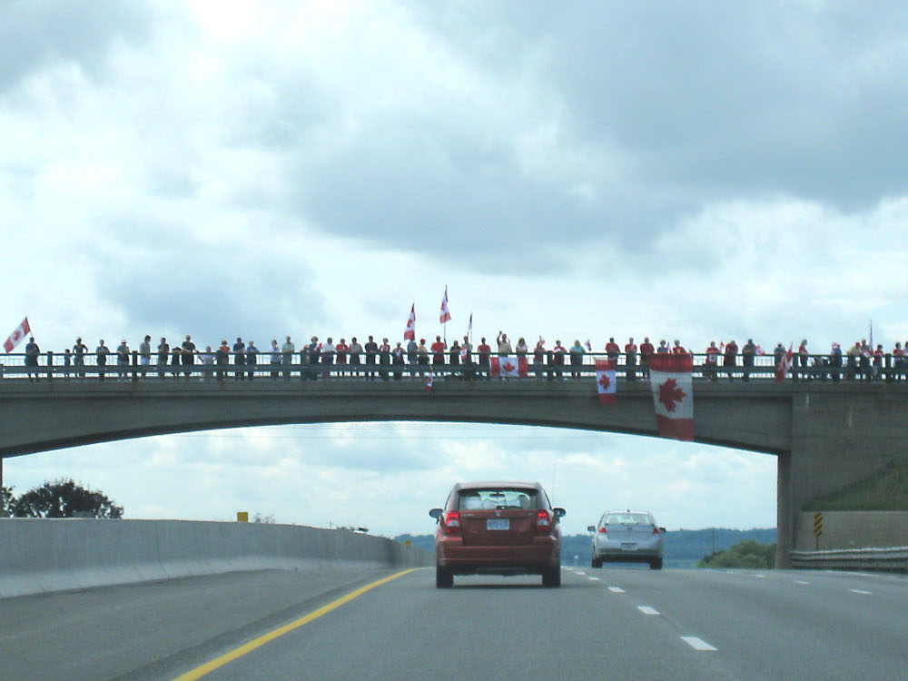 Des voitures circulent sur une autoroute, sous un viaduc bondé de gens, dont certains brandissent de grands drapeaux canadiens.