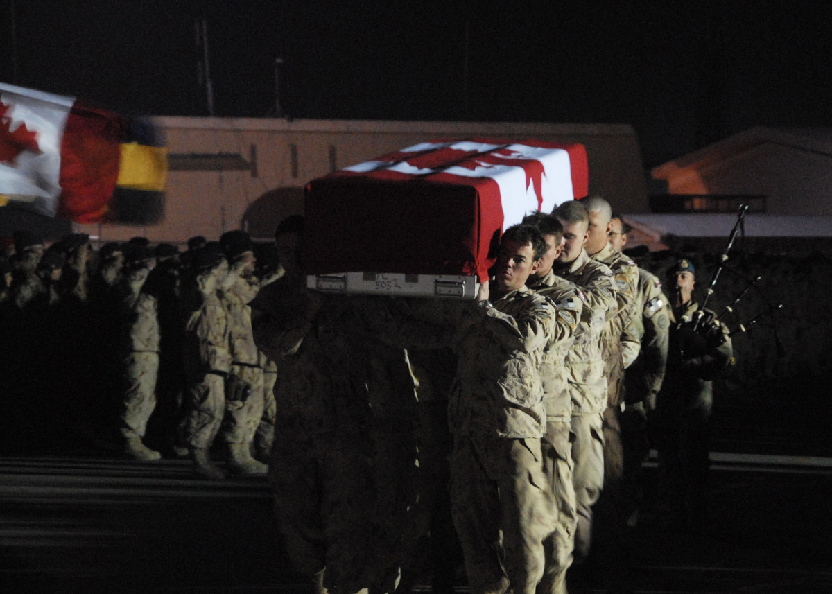 De nuit, sous un éclairage vif venant de la droite, huit militaires en uniforme beige s’avancent vers la caméra, portant sur leurs épaules une boite rectangulaire recouverte d’un grand drapeau canadien; derrière se trouvent un joueur de cornemuse, d’autres militaires et un autre drapeau.