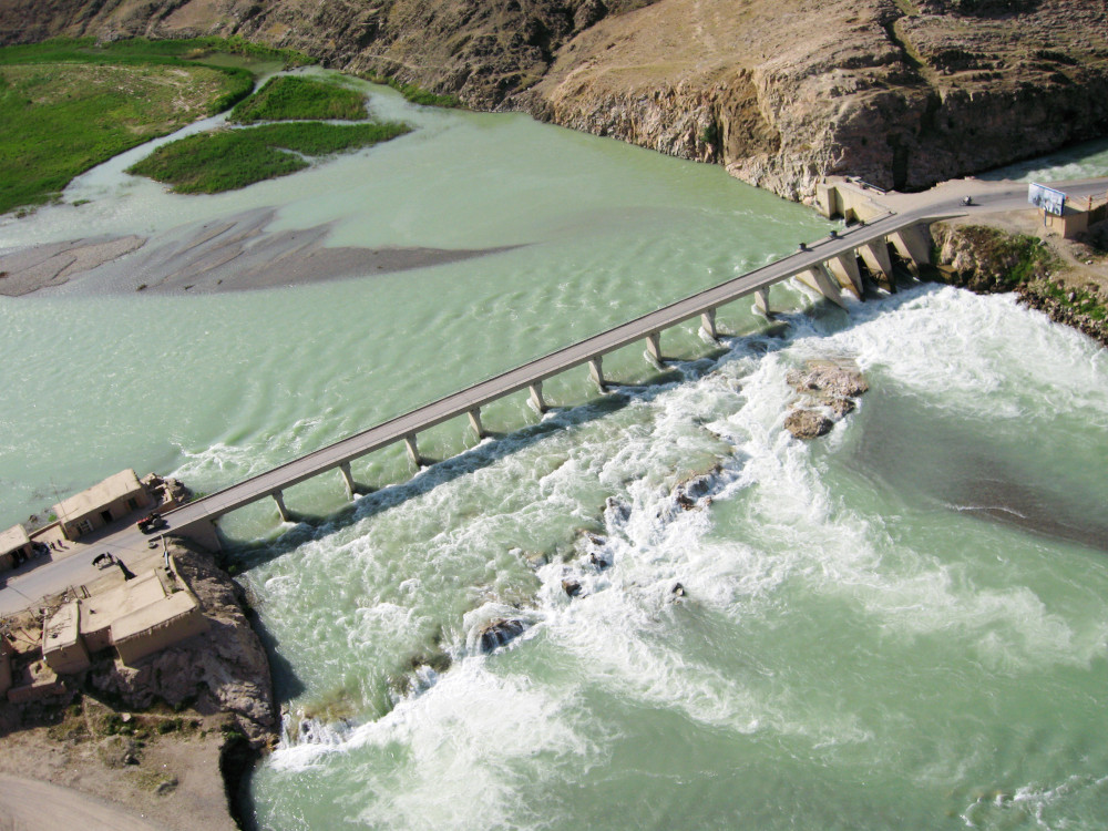 Un pont étroit enjambe un large cours d’eau bleu-vert qui s’écoule par-dessus de faibles rapides.