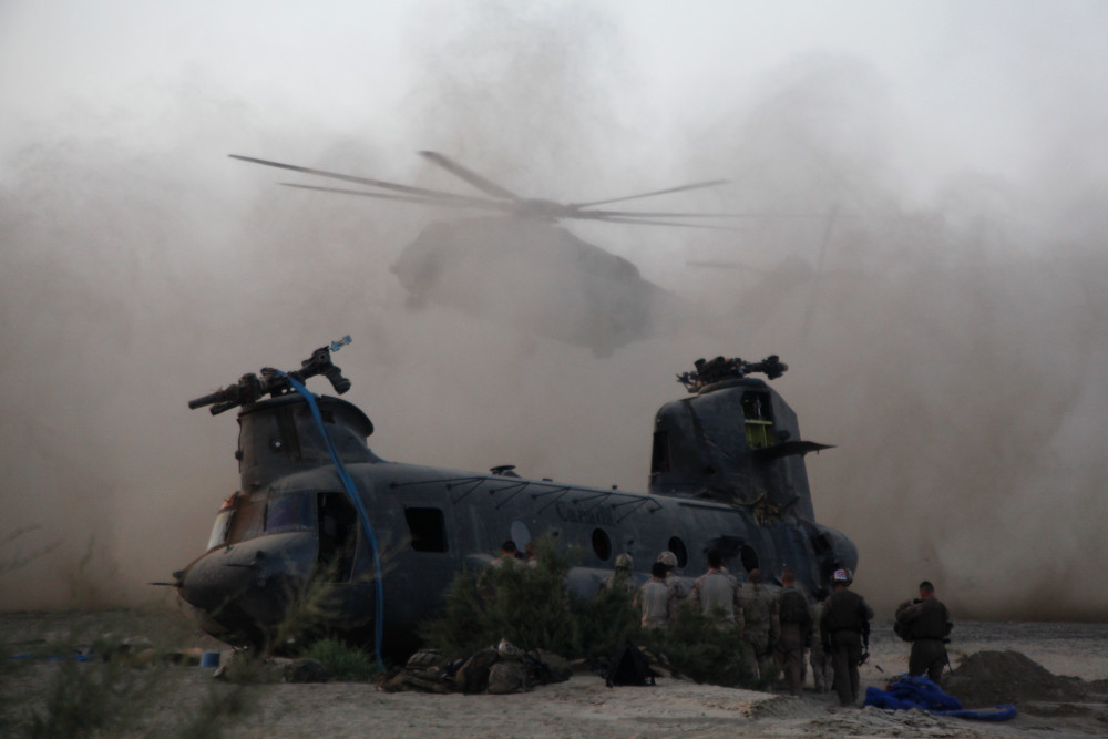 Un groupe de militaires devant un gros hélicoptère; un autre hélicoptère atterrit en arrière-plan dans un énorme nuage de poussière.