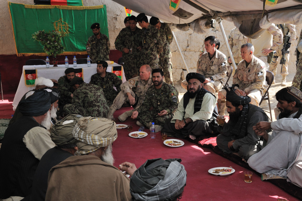 Un groupe d’hommes, vêtus à la fois d’uniformes militaires occidentaux et de robes traditionnelles afghanes, est assis sur un tapis rouge; plusieurs d’entre eux ont devant eux des assiettes de nourriture et des verres.