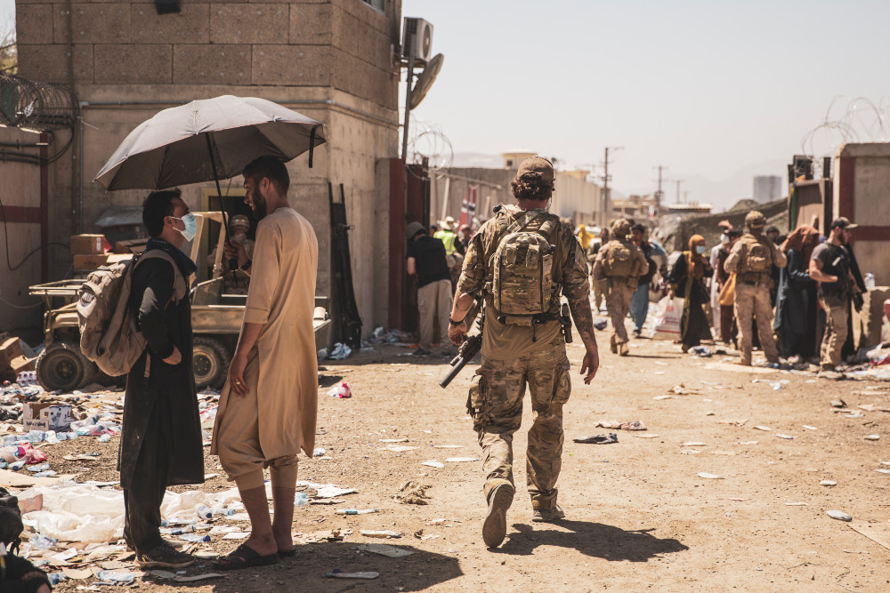 Un soldat en tenue de camouflage s’avance vers une foule nombreuse de militaires et de personnes civiles, au milieu de murs surmontés de fil barbelé.