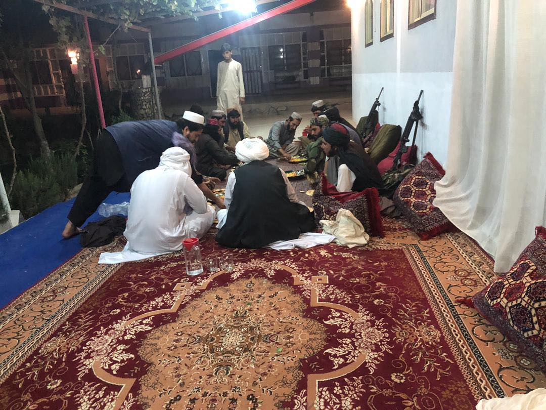 Un groupe de personnes au teint moyen, vêtues de vêtements amples et portant des voiles, est assis sur une terrasse recouverte de tapis, près d’une rangée d’oreillers et d’armes automatiques appuyées contre un mur.