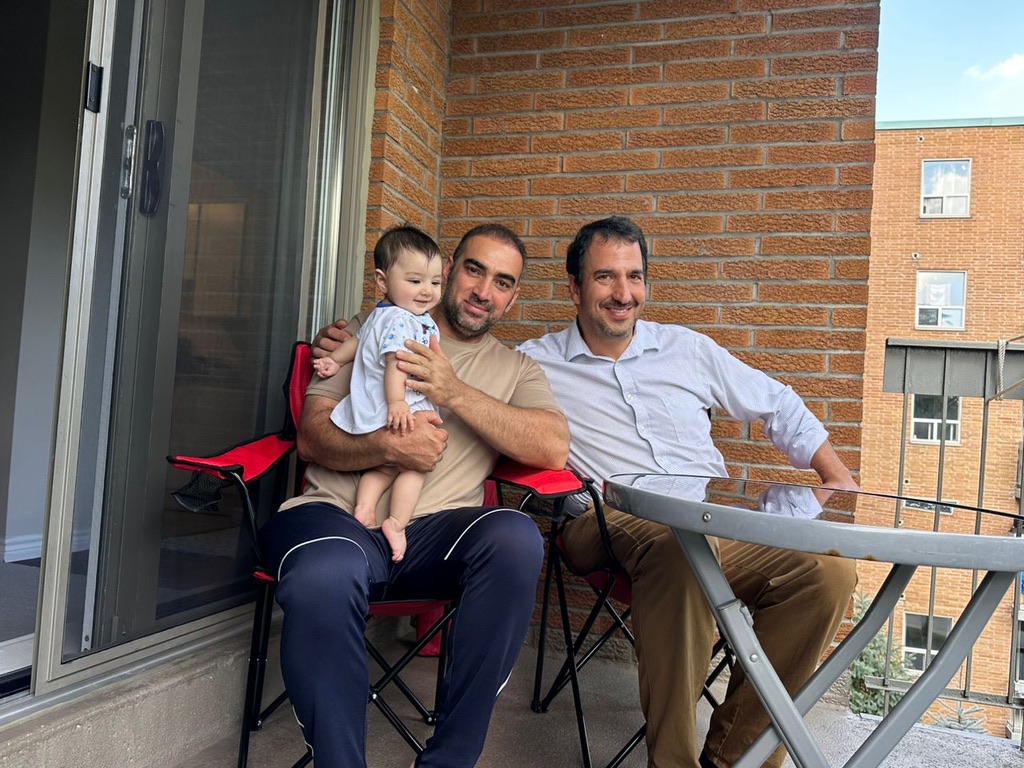 Un homme au teint moyen, souriant, tient un bébé dans ses bras et est assis à côté d’un homme au teint pâle, lui aussi souriant, sur le balcon d’un appartement.