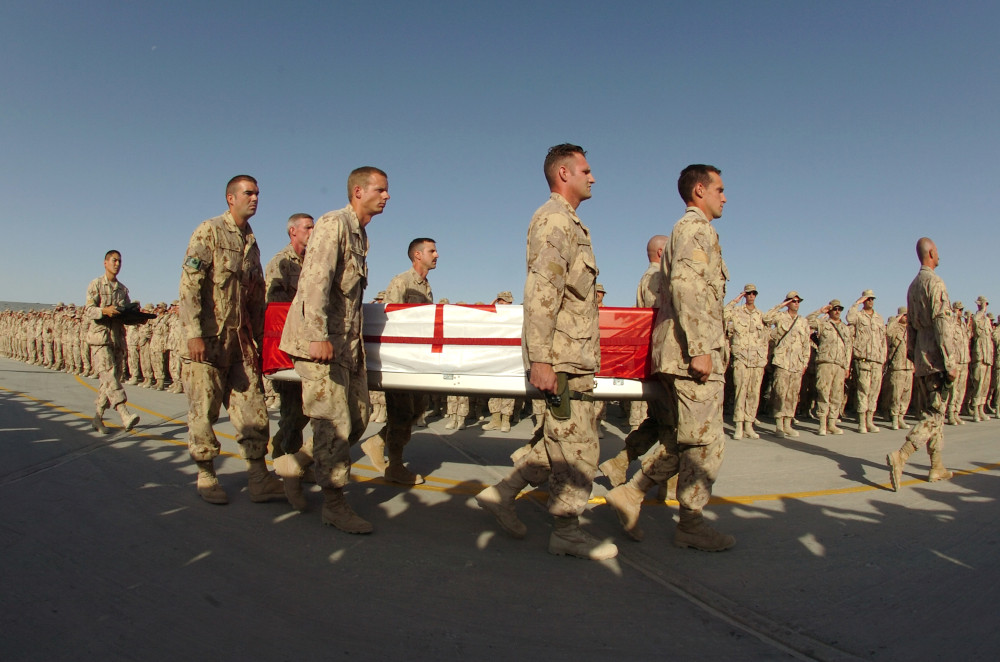 Un cortège de militaires en tenue de camouflage beige transporte un cercueil métallique [HR1.1]recouvert du drapeau canadien devant une longue file de militaires qui saluent.