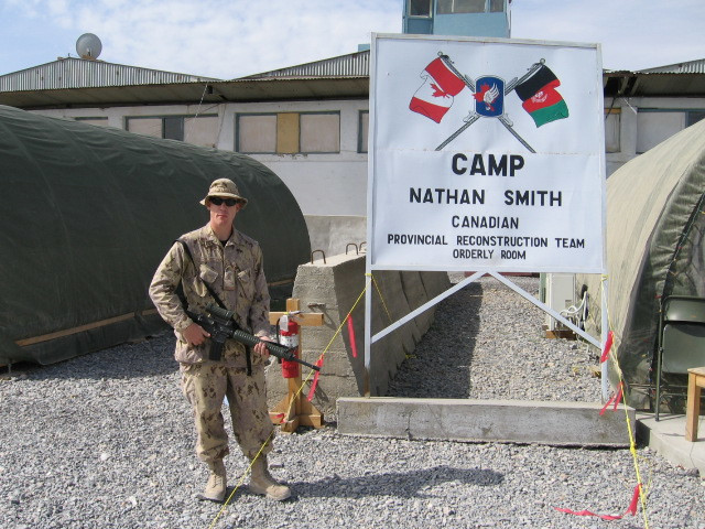 Un soldat vêtu d’un uniforme beige et armé se tient près d’un grand panneau blanc sur lequel figurent les drapeaux canadien et afghan entrecroisés, avec l’inscription « Équipe provinciale de reconstruction canadienne du Camp Nathan Smith » [traduction].