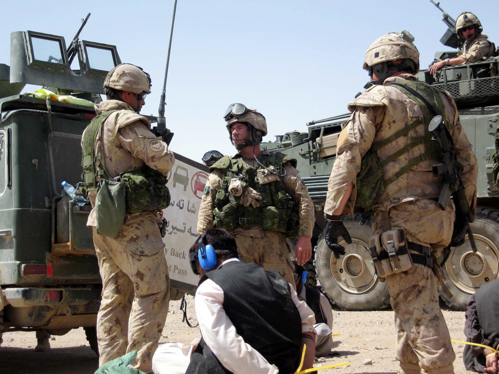Trois militaires avec leur arme, en tenue de camouflage beige, près de deux gros véhicules blindés verts, avec des [HR7.1]personnes en civil assises par terre, les yeux bandés, portant des protège-oreilles en plastique bleu et les mains liées derrière le dos.