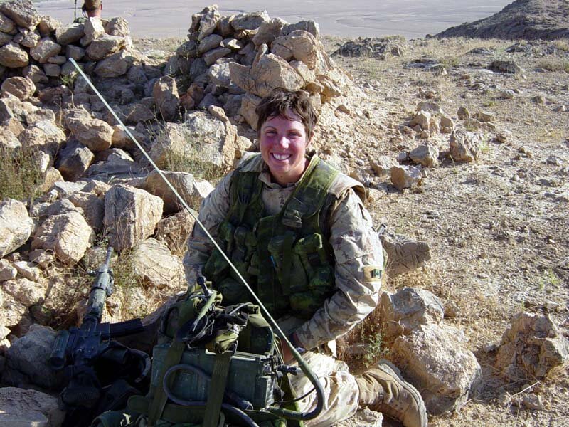 Une soldate souriante, aux cheveux courts et ondulés, vêtue d’une tenue de camouflage beige et d’un gilet pare-balles vert, est assise dans un paysage rocheux.