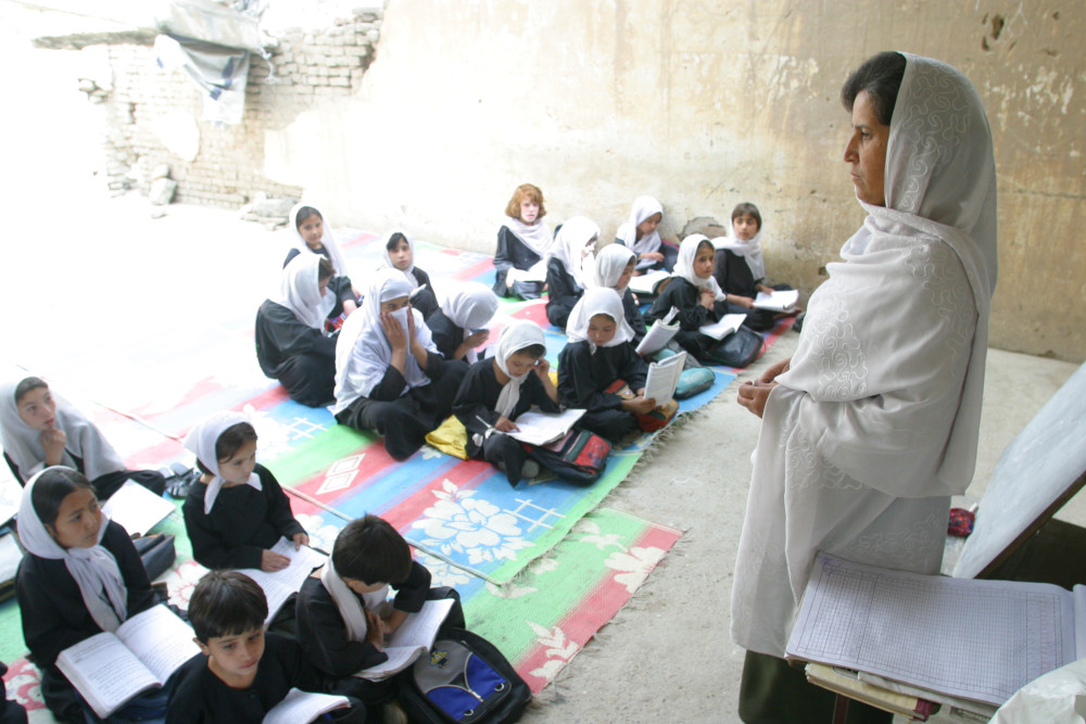Une enseignante portant un long voile s’adressant à une classe d’enfants prenant place par terre, dont la plupart portent aussi un voile.