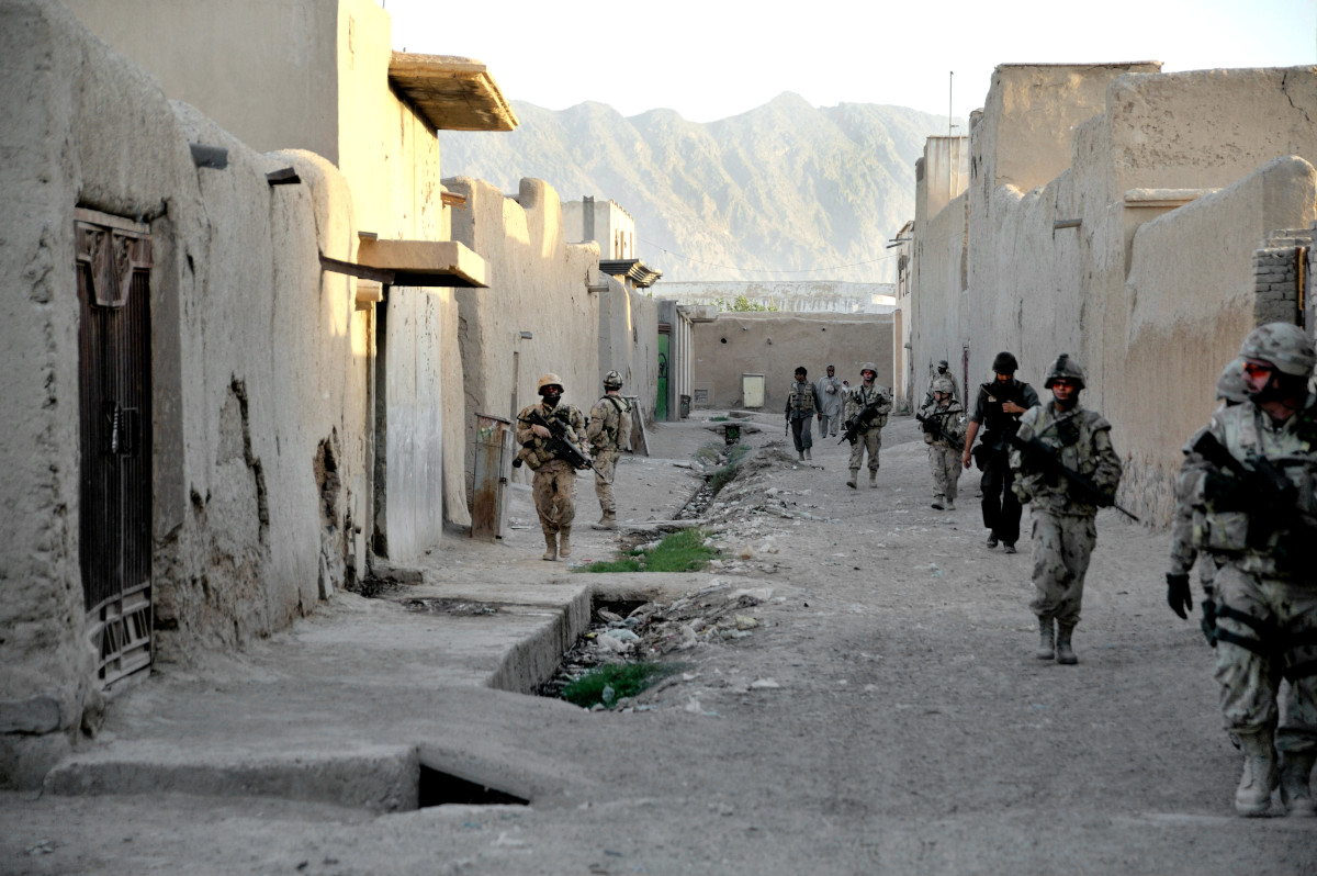 Plusieurs militaires avec leurs armes marchant de part et d’autre d’une rue bordée de bâtiments bas et de murs.