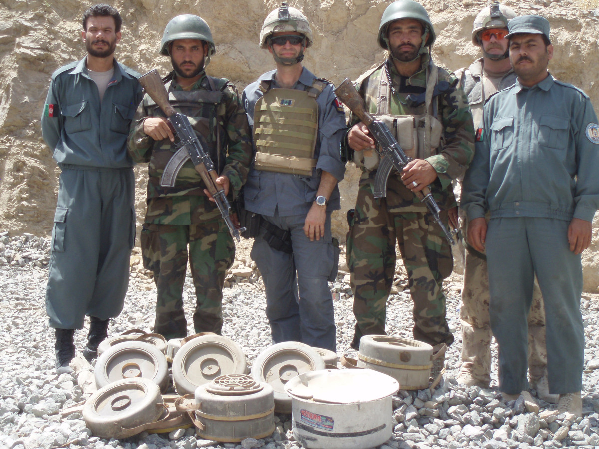 Six personnes, certaines vêtues d’uniformes bleus, d’autres d’une tenue de camouflage vert ou beige clair, se tiennent devant une dizaine d’objets cylindriques de petite taille, parmi lesquels des mines terrestres et des pots
