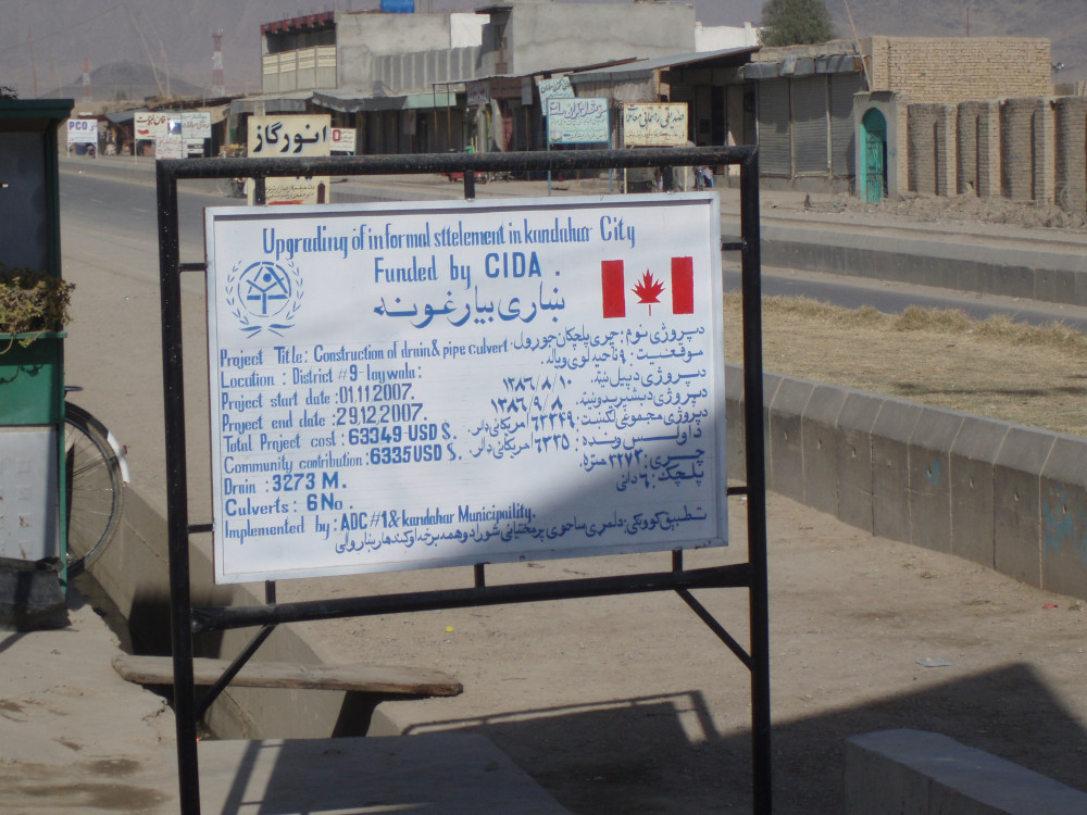 Un panneau routier rédigé en anglais et en pachto, sur lequel figure en gros caractères le titre « Amélioration d’un quartier informel de la ville de Kandahar financée par l’ACDI », au-dessus d’une liste de renseignements présentés en caractères beaucoup plus petits