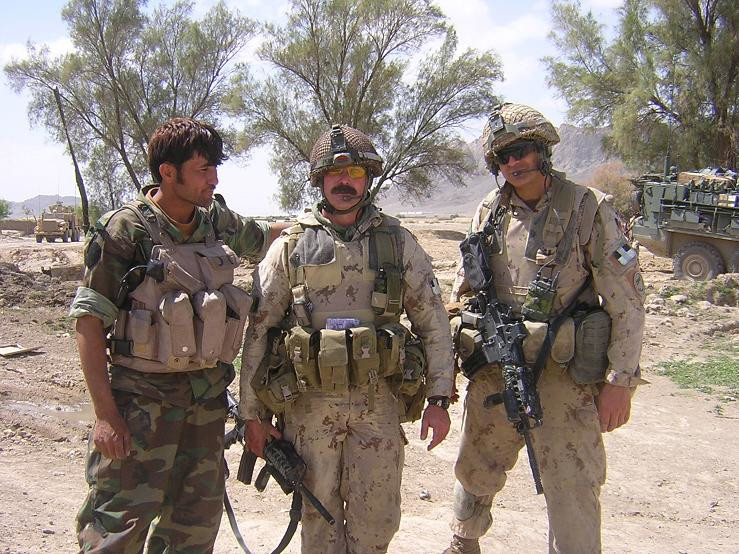 Un soldat à la peau moyennement foncée, vêtu d’une tenue de camouflage vert, se tenant à côté de deux soldats à la peau pâle, vêtus d’une tenue de camouflage beige; on aperçoit des arbres et des véhicules blindés en arrière-plan.