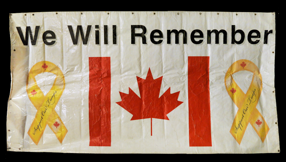 Une bannière rectangulaire en vinyle représentant un drapeau canadien flanqué de deux rubans jaunes portant l’inscription « Support Our Troops (Soutenons nos troupes[HR4.1]) », sous l’inscription en gros caractères « We Will Remember (Nous nous souviendrons) ».